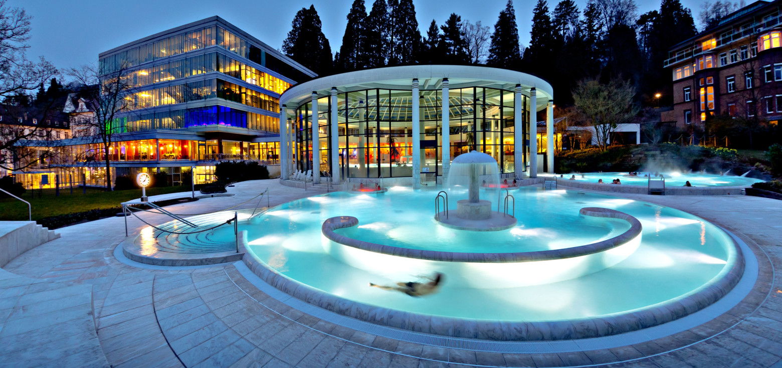Beleuchtetes Außenbecken der Caracalla Therme Baden-Baden in der Abenddämmerung mit warmem Thermalwasser, Springbrunnen und moderner Glasarchitektur im Hintergrund.