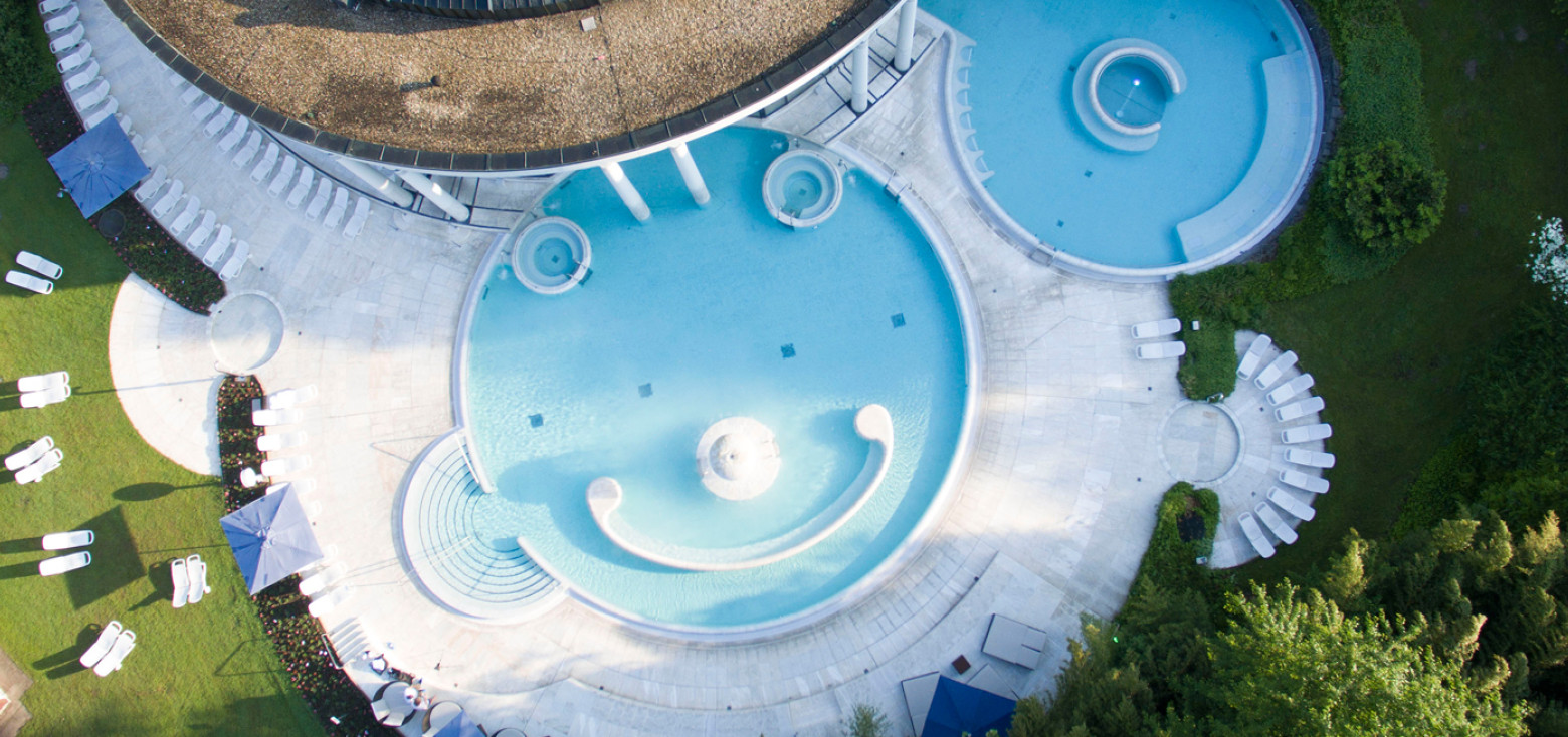 Drohnenaufnahme des runden Außenbeckens der Caracalla Therme in Baden-Baden mit hellblauen Fliesen, Sitzbereichen im Wasser und umliegender Liegewiese.