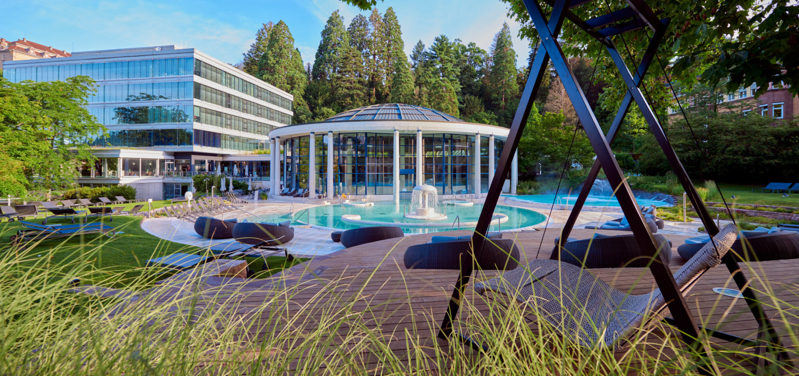 Caracalla Therme Baden-Baden mit Glasrotunde und Außenbecken, fotografiert durch Gräser im Vordergrund.