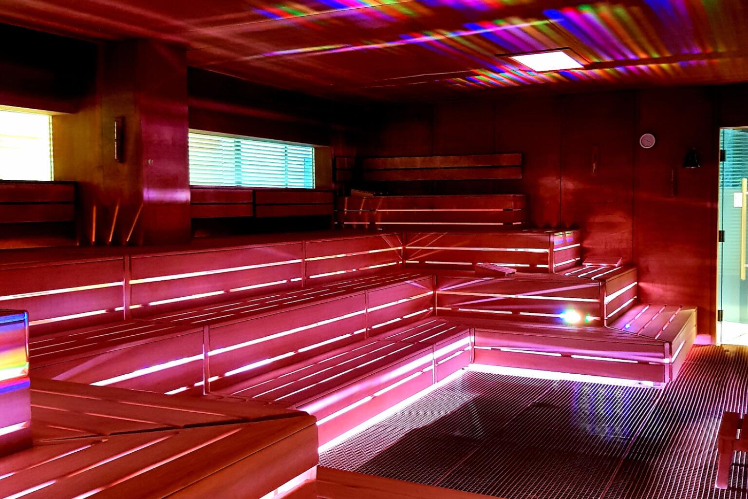 Innenansicht der Spectaculum Sauna – ein großer würfelförmiger Raum mit gestaffelten Holzbänken. Bunte Lichteffekte in Rot, Blau, Gelb und Grün brechen sich an den Wänden und Bänken, wodurch ein farbiges Spiel entsteht.