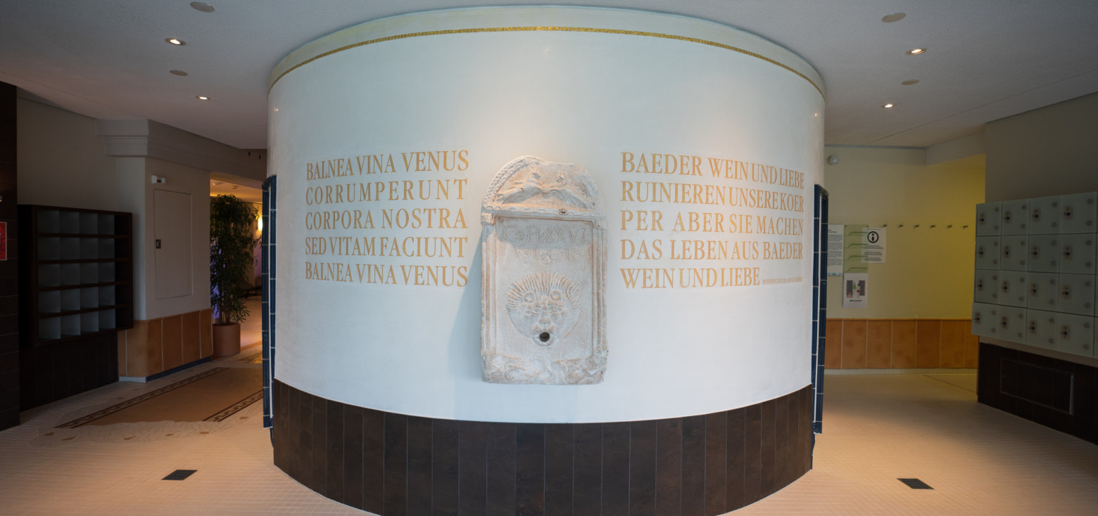 Rundwand im Eingangsbereich der Therme mit römischem Wandbrunnen aus Stein und goldfarbenen lateinischen sowie deutschen Inschriften.